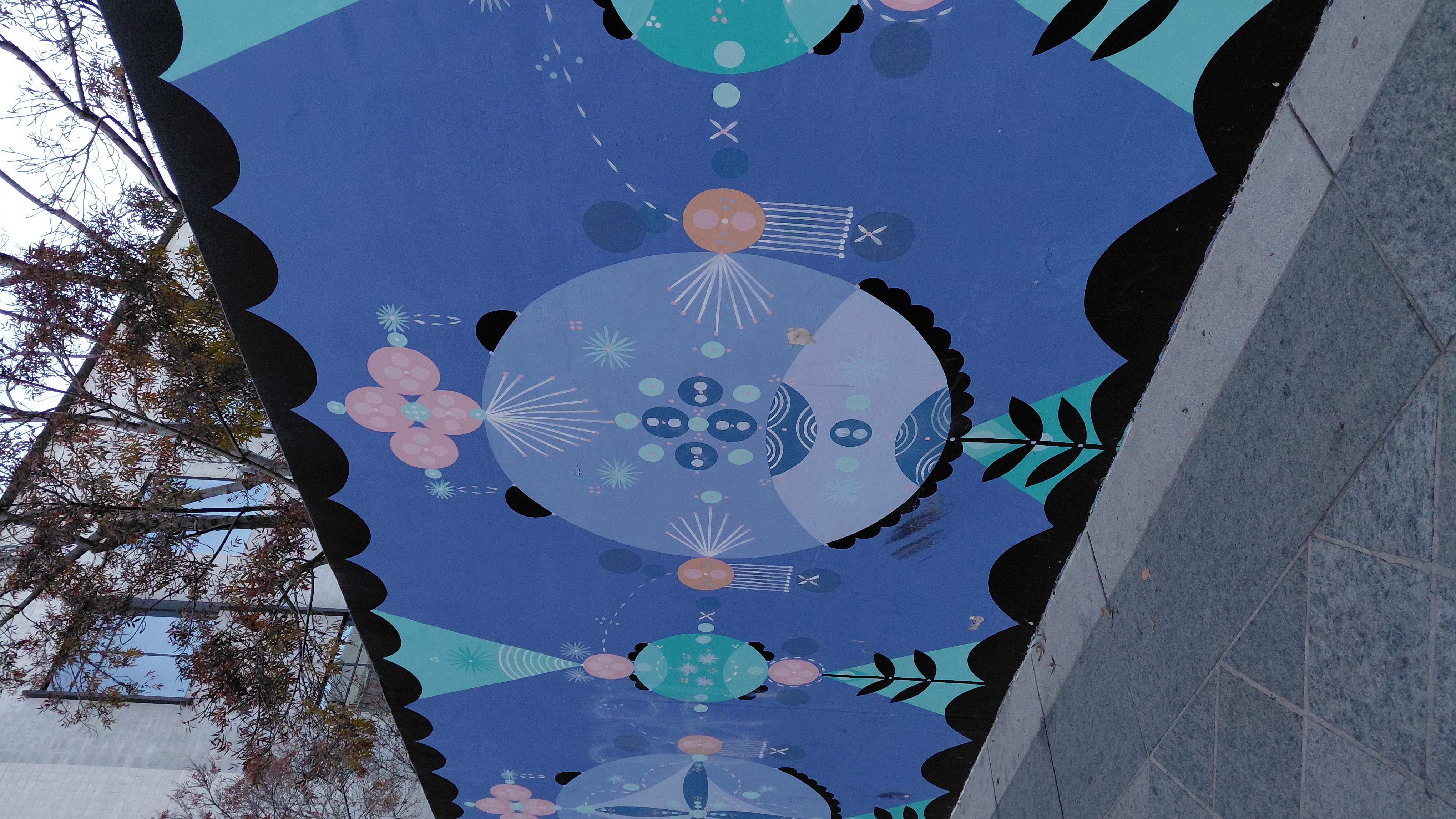 Long blue wall with geometric mandala-style mural along a sidewalk